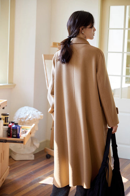 Full-length back view of almond-camel cashmere wool long coat as model walks away in studio with art supplies