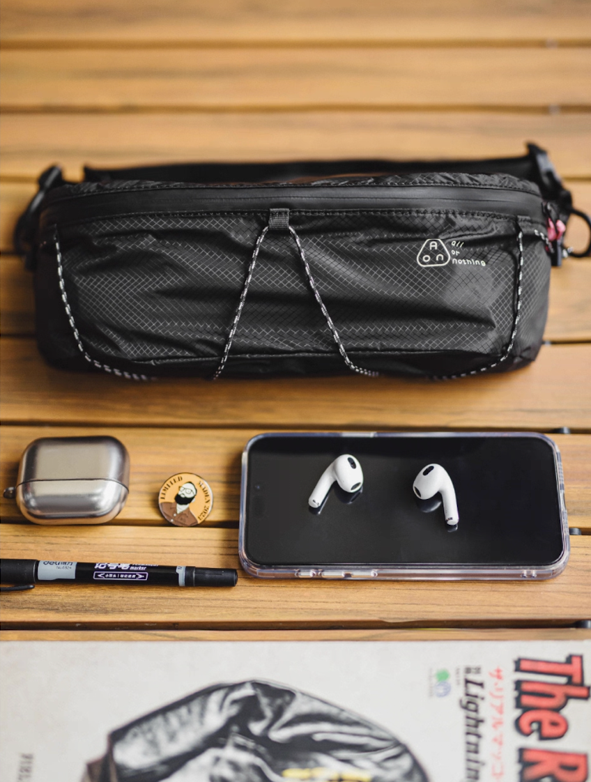 Black techwear sling bag on wood surface with phone and AirPods