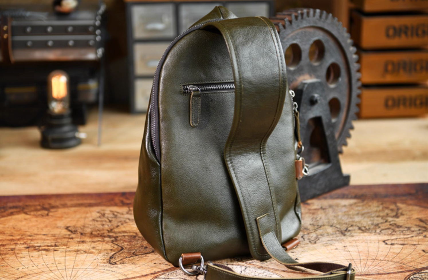 Rear view of army green leather sling bag with zipper pocket and wide strap resting on wooden surface.