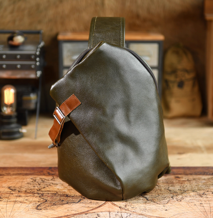 Front view of army green full-grain leather sling bag with tan buckle strap on wooden table background.