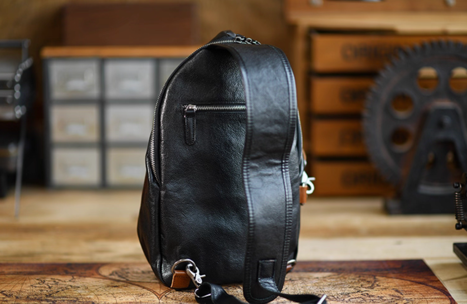 Rear view of black leather sling bag with zipper pocket and wide shoulder strap on wooden table.