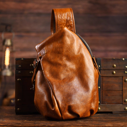 Hand-dyed caramel brown leather sling bag standing on wooden chest, front view.