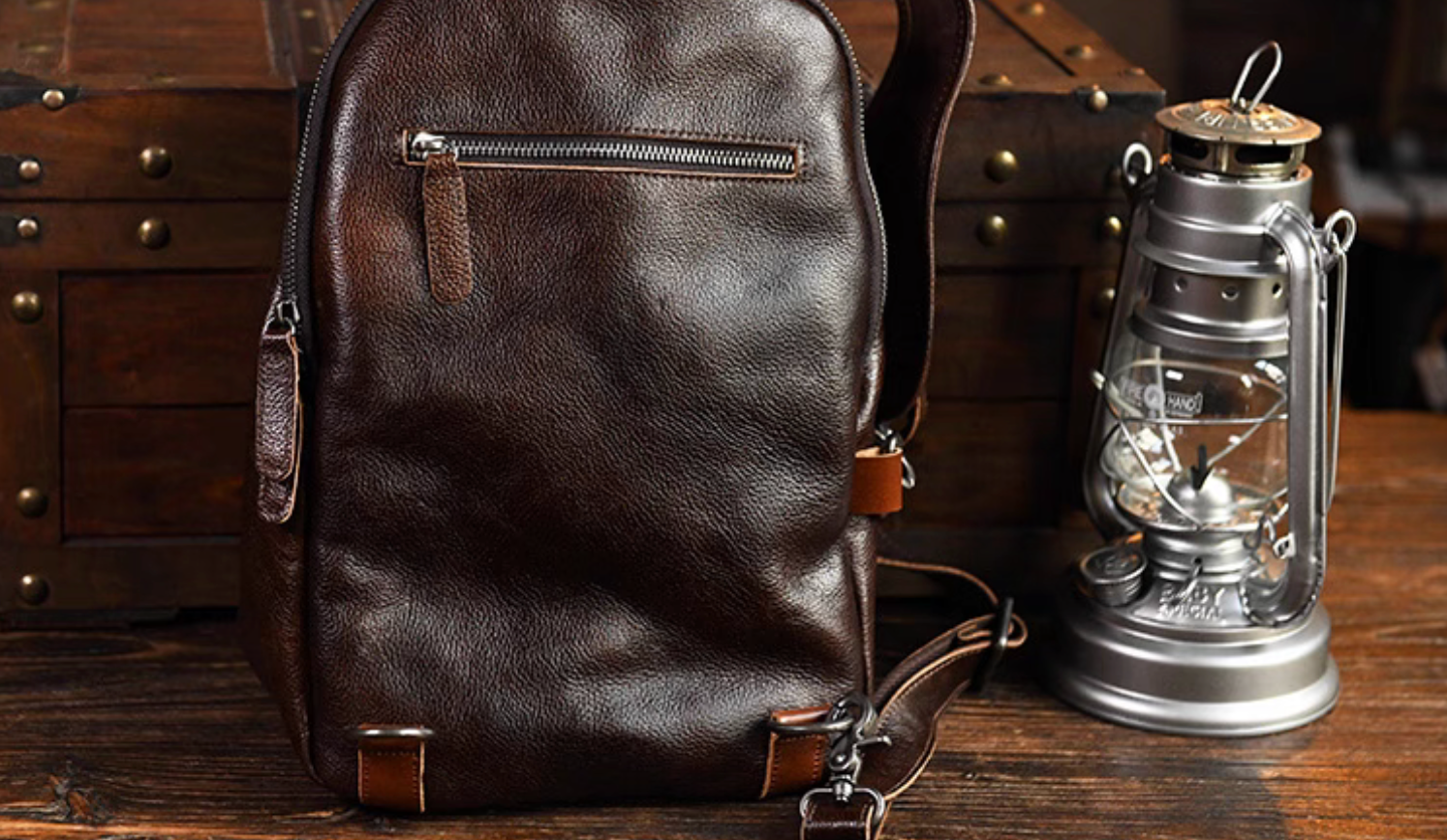 Back view of dark brown leather sling bag with rear zipper pocket beside vintage lantern and wooden chest.