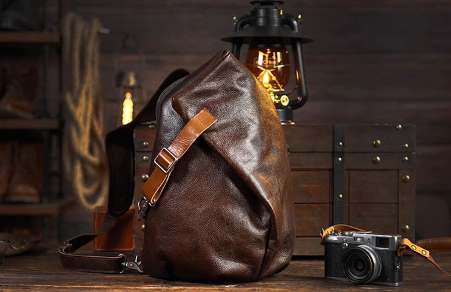 Dark brown leather sling bag on rustic table with lantern, showing rear zipper pocket.