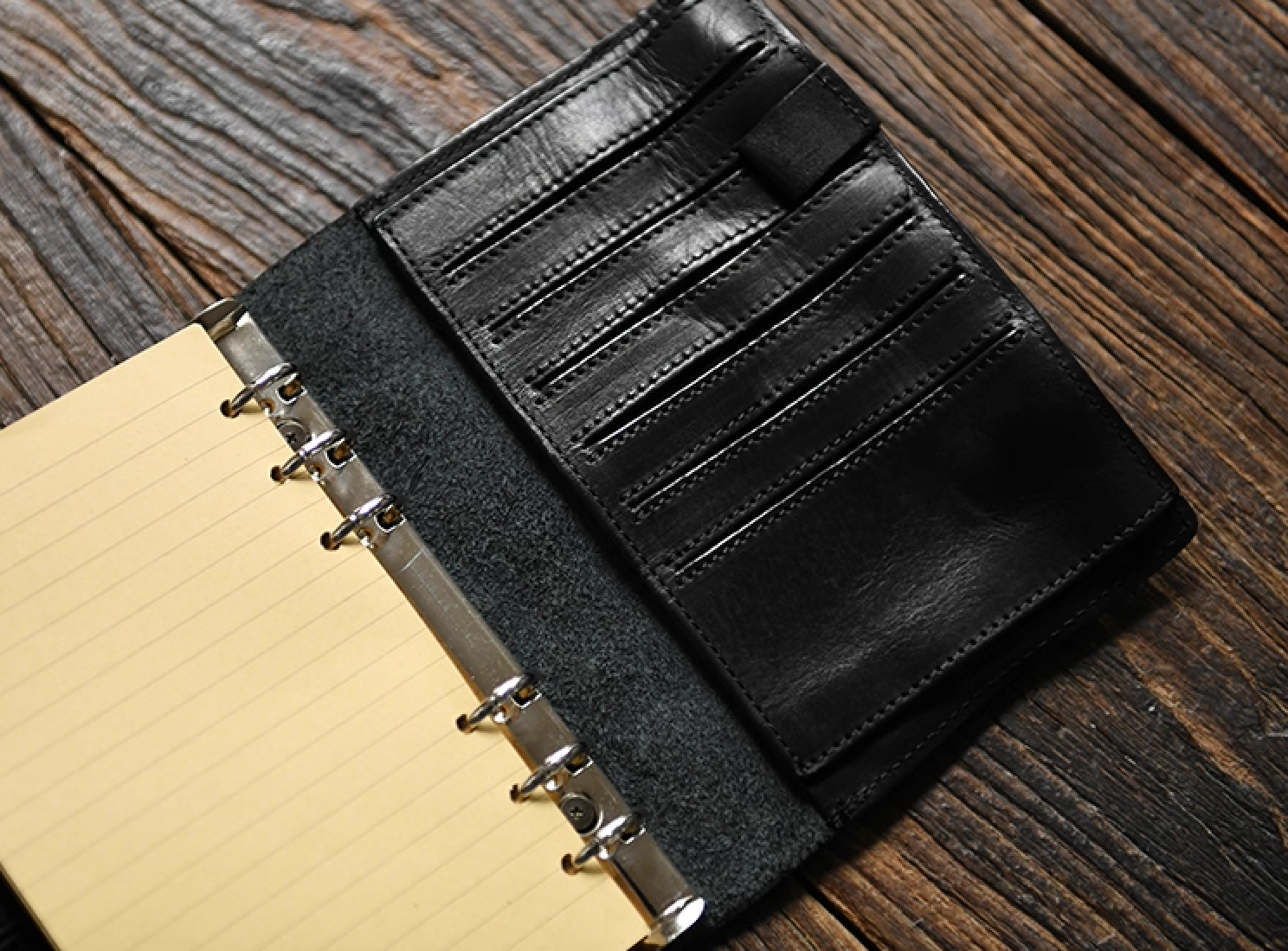 Open black leather refillable journal on rustic wood table showing ring binder, yellow lined pages, and multiple leather card slots.