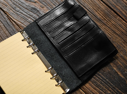 Open black leather refillable journal on rustic wood table showing ring binder, yellow lined pages, and multiple leather card slots.