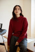 Model wearing red Bordeaux Wool-Cashmere Turtleneck Sweater sitting on desk showcasing relaxed fit, ribbed hem, and soft knit texture
