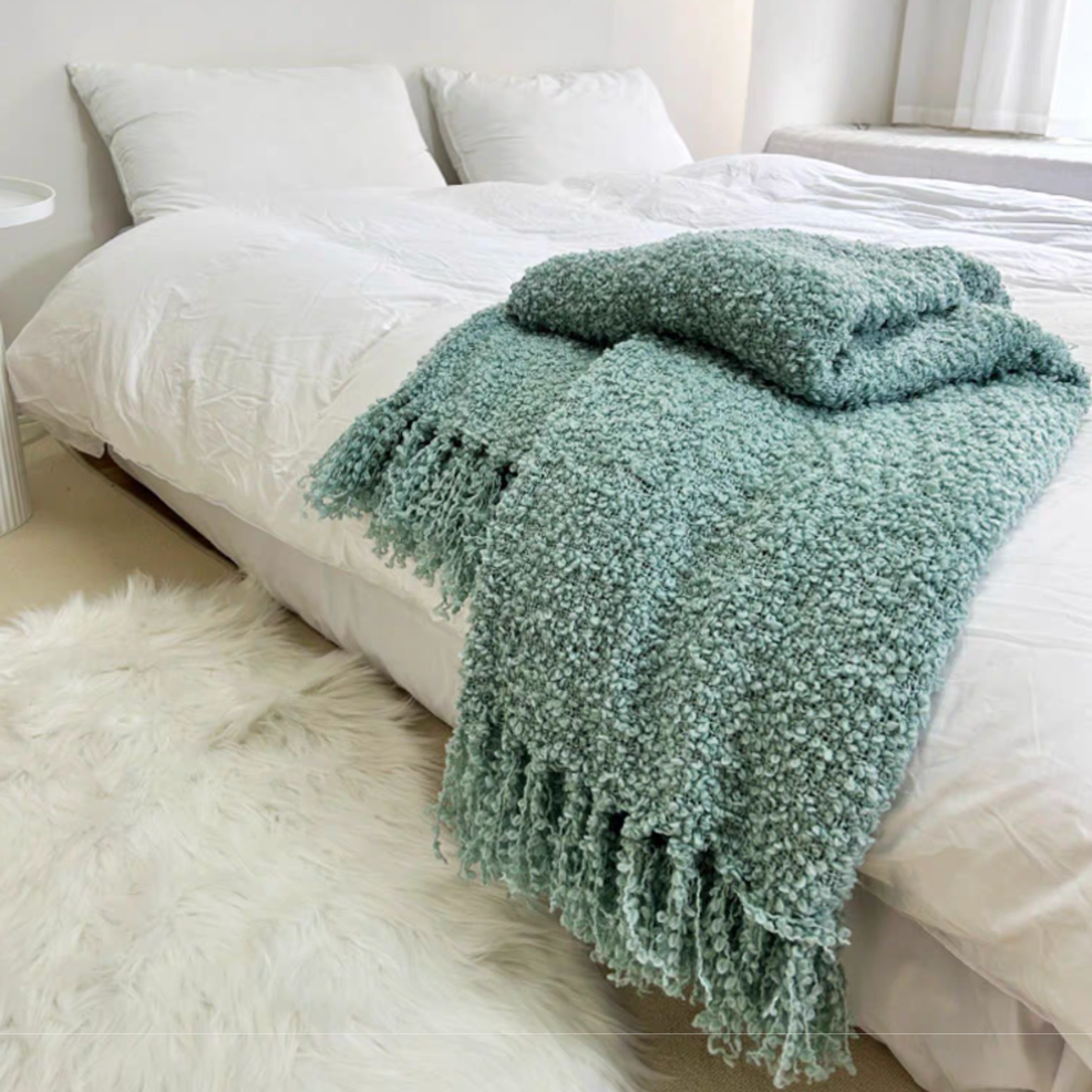 Blue textured loop knit throw blanket with tassel fringe folded on white bed in minimal bedroom