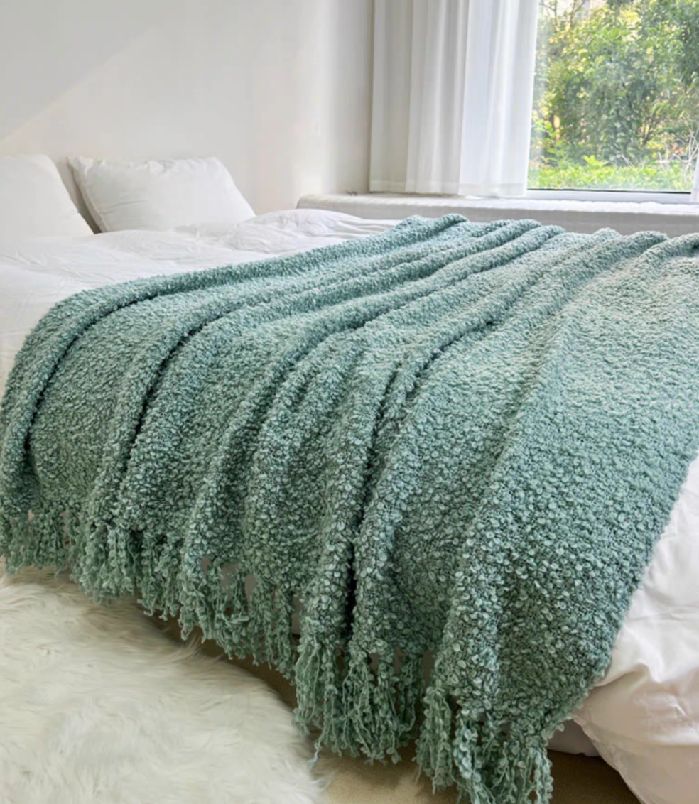 Blue loop knit throw blanket with tassel fringe fully draped across bed with natural window light