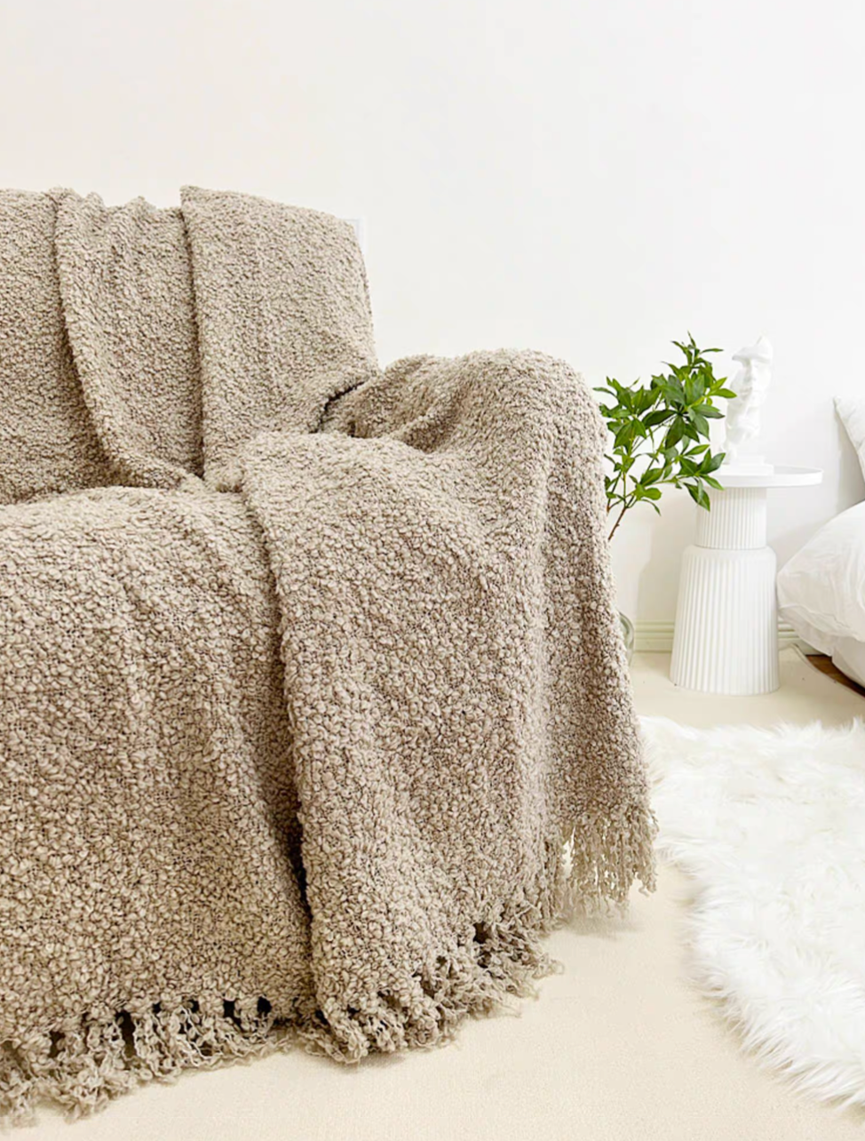 Gray loop knit throw blanket with tassel fringe draped over armchair in bright minimal room