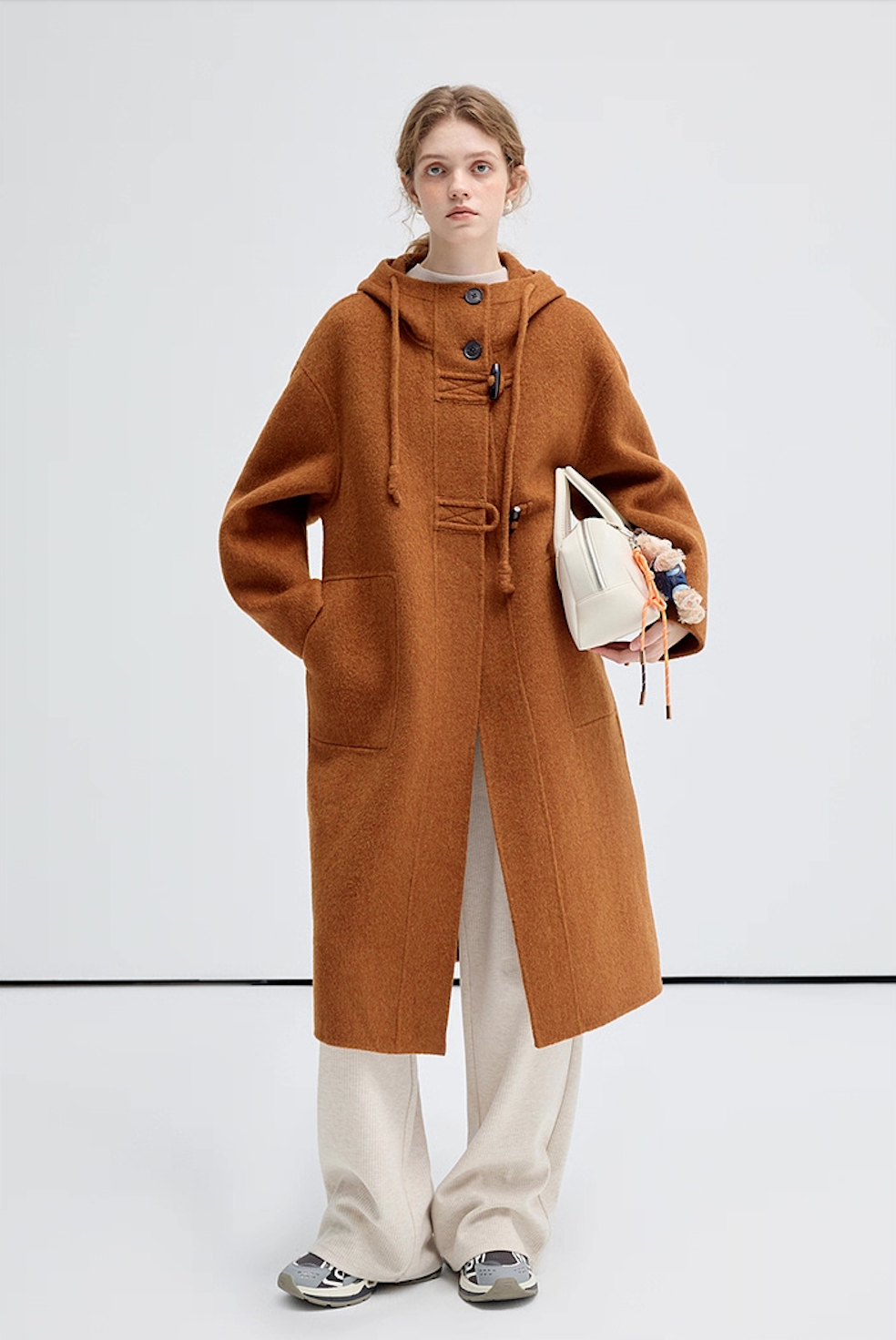 Model wearing a brown hooded wool coat, holding a white handbag, full front view.
