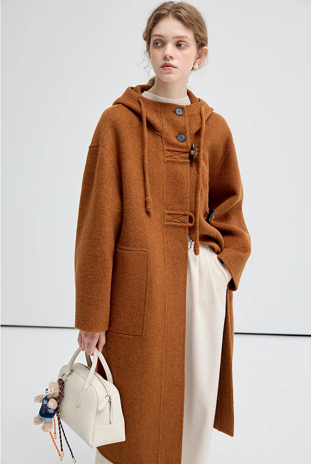 Female model in brown hooded coat, half-body view with white handbag.