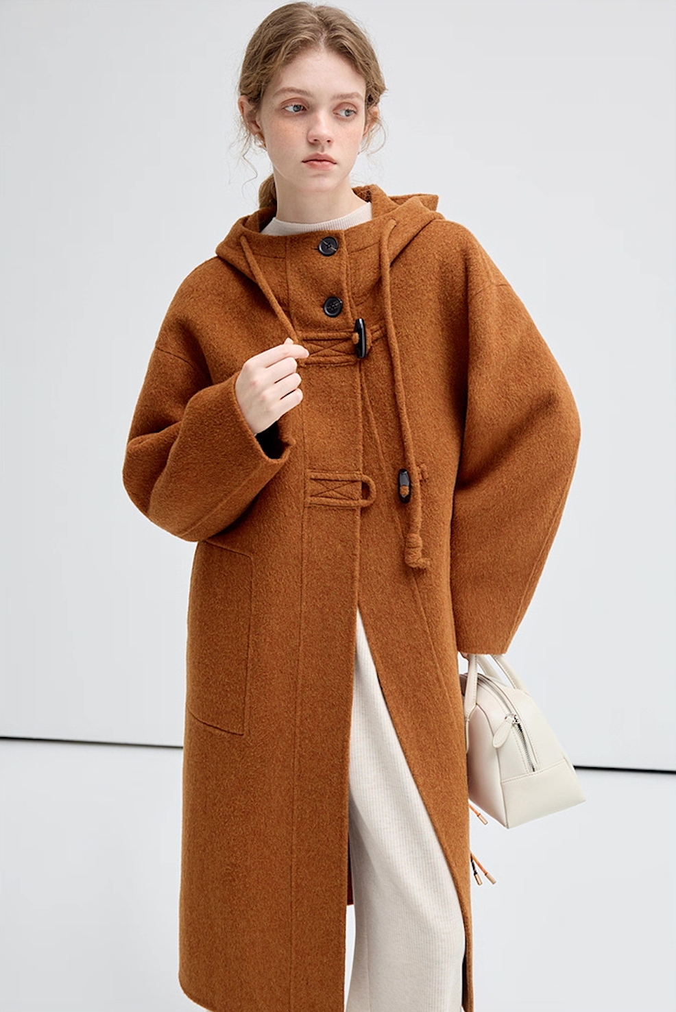 Model wearing brown hooded wool coat, half-body pose with white handbag, front view.