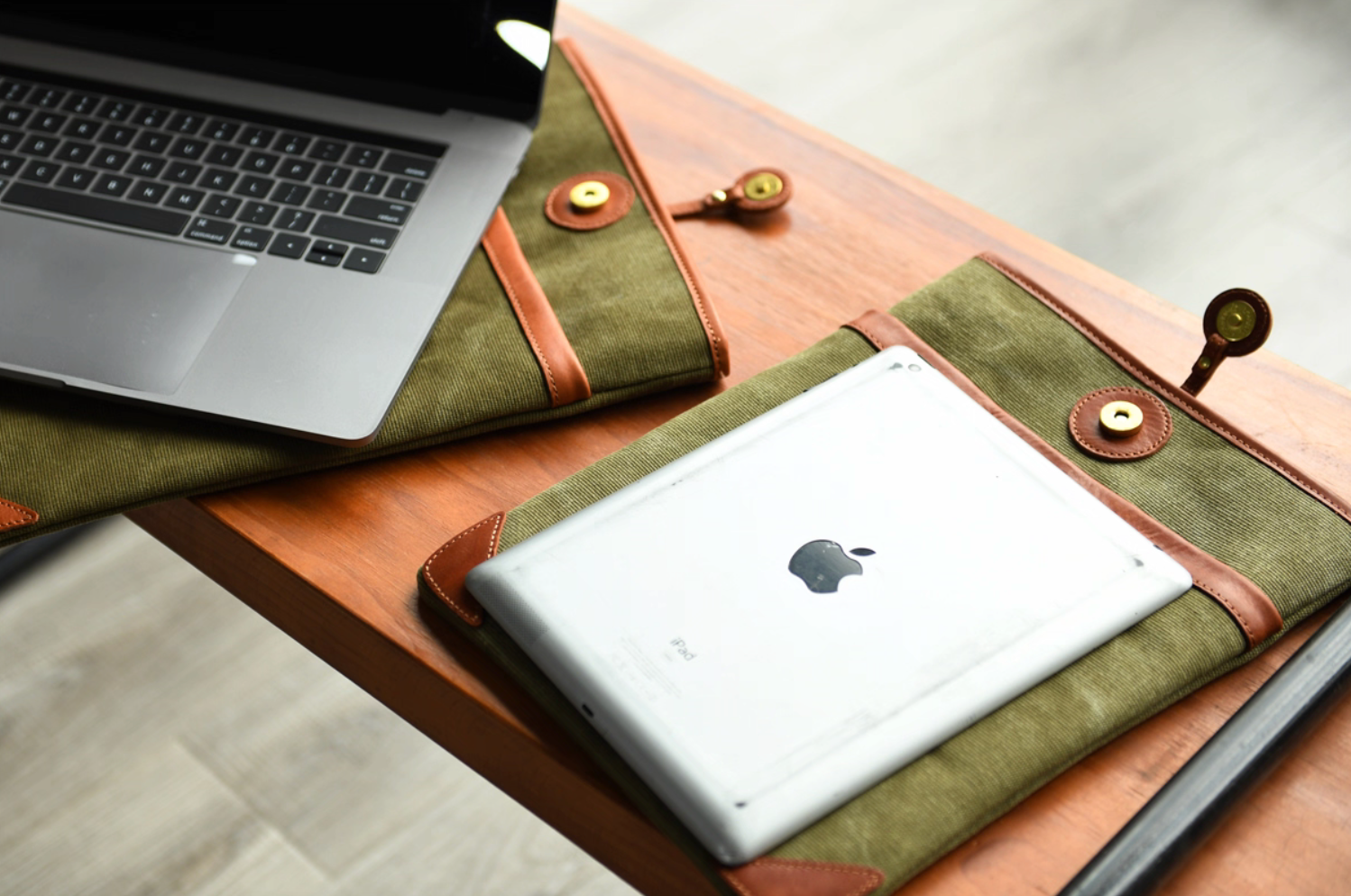 Olive green canvas and leather laptop sleeves on a desk with a MacBook and iPad partially inside the sleeves.