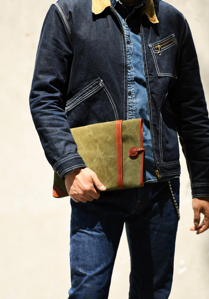 Man in denim jacket holding an olive green canvas laptop sleeve with brown leather trim outdoors.