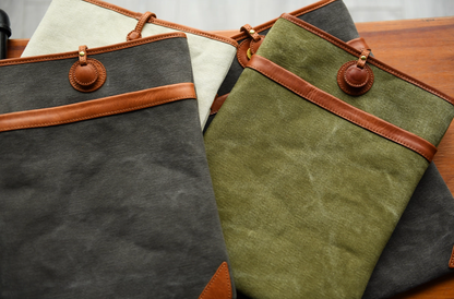 Canvas and leather laptop sleeves in charcoal, ivory, and olive with tan leather trim arranged on a wooden surface.
