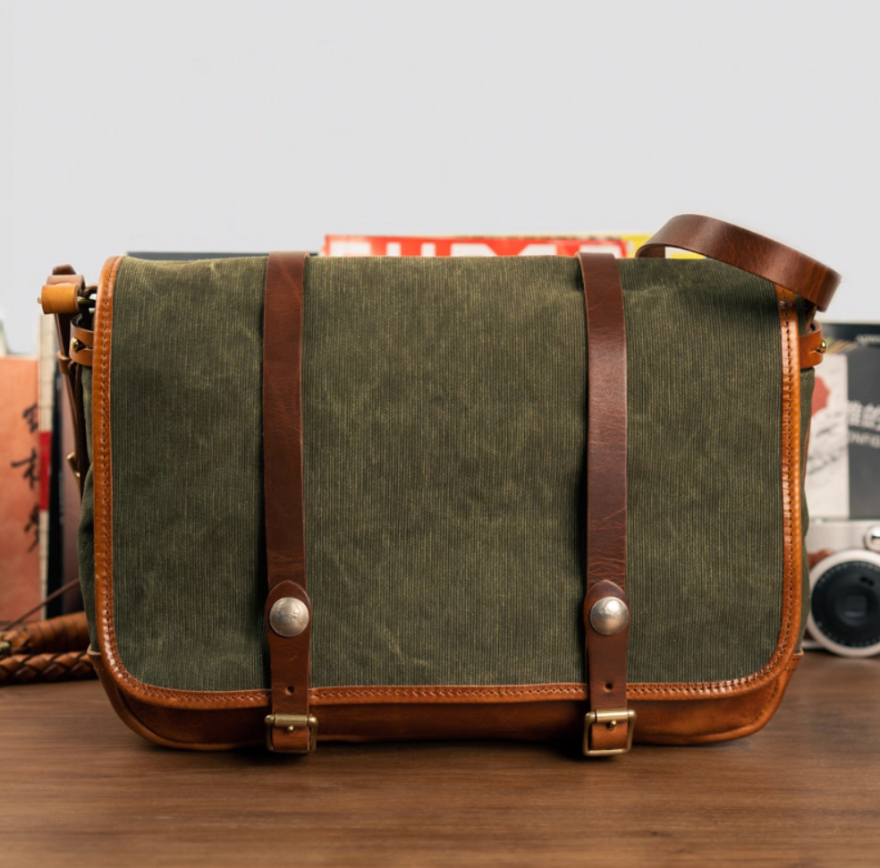 Front view of army green canvas messenger bag with brown leather trim and buckle straps on desk
