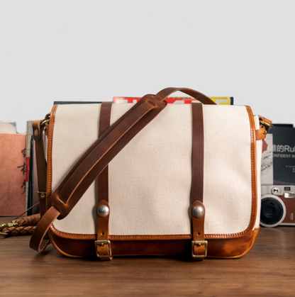 Front view of beige canvas and tan leather messenger bag with buckle straps on desk