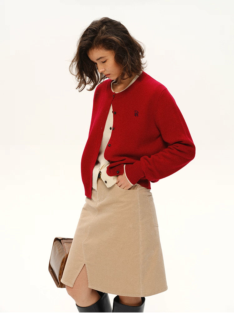 Model wearing cherry red wool logo cardigan styled with beige skirt, hands in pockets, front profile view