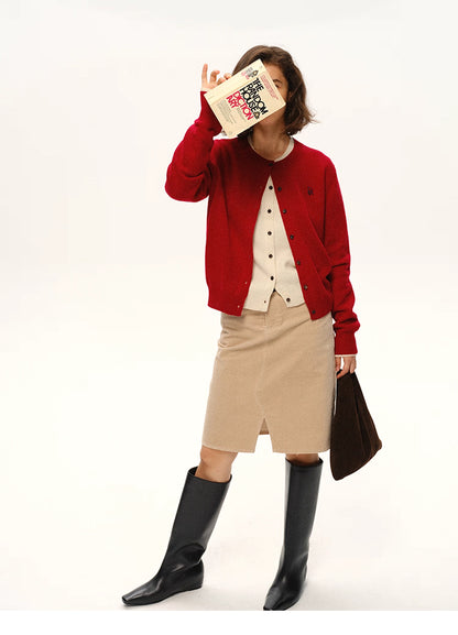 Model wearing cherry red wool cardigan holding a book in front of face, styled with beige skirt and black boots