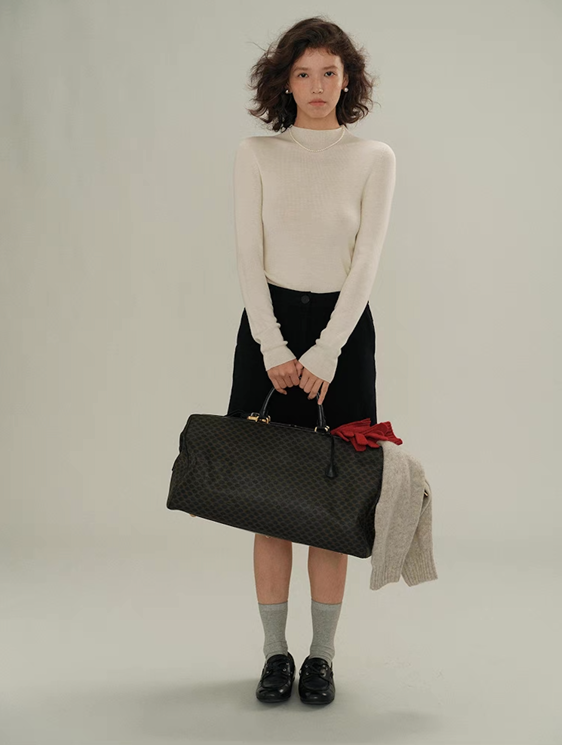 Full-body shot of woman in white cashmere sweater and black skirt holding a travel bag – elegant minimalist fashion.