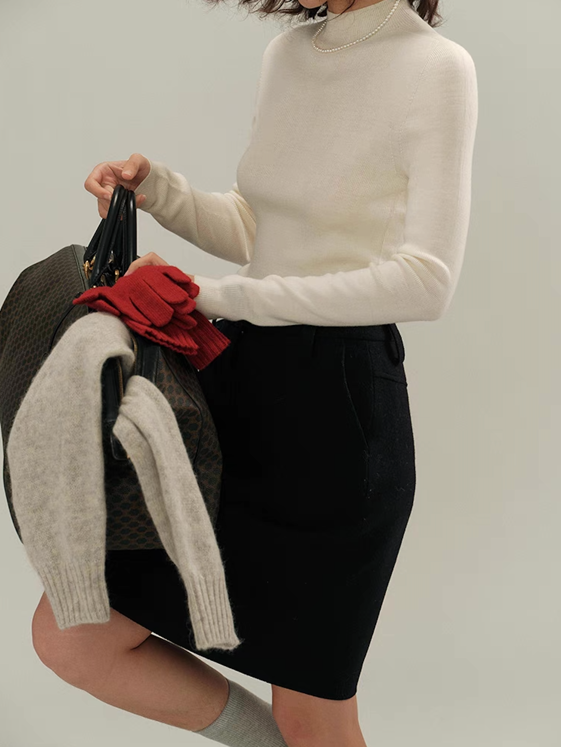 Woman in ivory white cashmere sweater with black skirt holding a bag and red gloves – chic winter look.