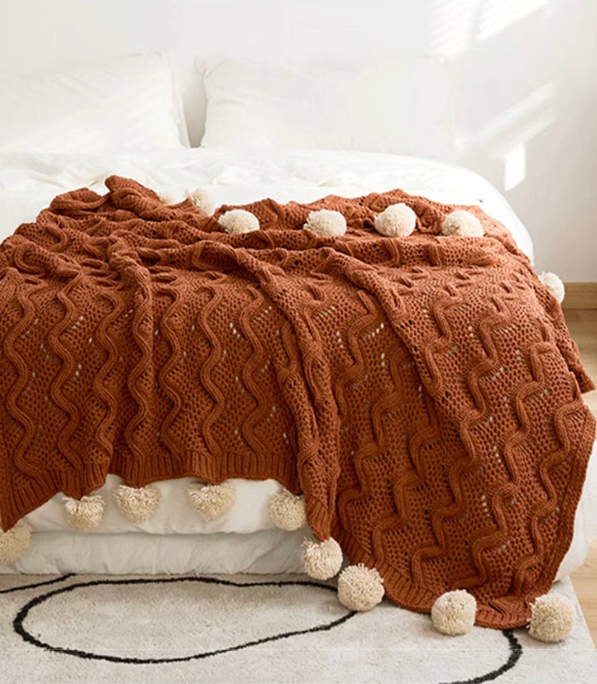 Burgundy chenille throw blanket layered across bed with textured knit pattern and pom pom edges