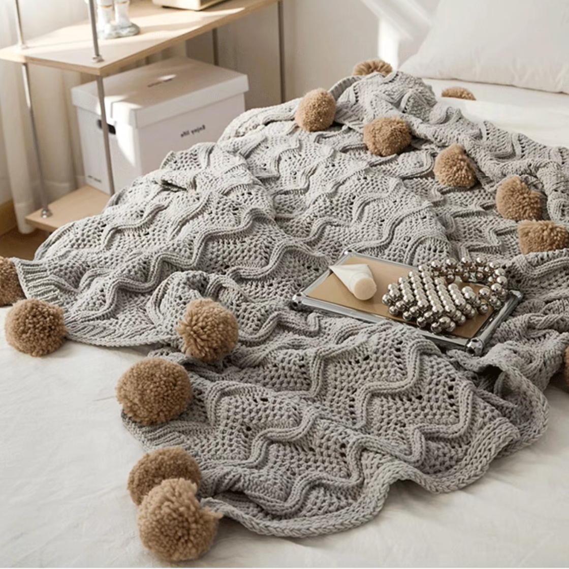Gray chenille throw blanket draped on bed with soft knit texture and pom pom tassels in cozy setting