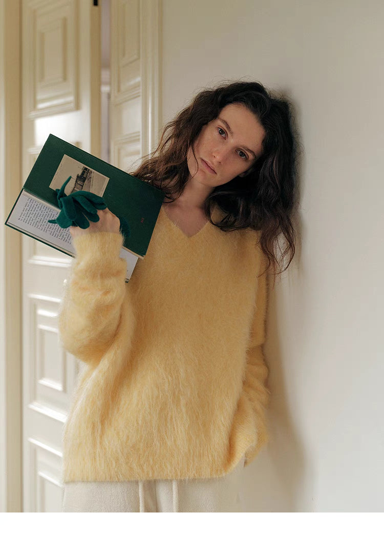 Cream yellow brushed wool V neck sweater leaning against wall holding book showcasing soft fuzzy texture and relaxed fit