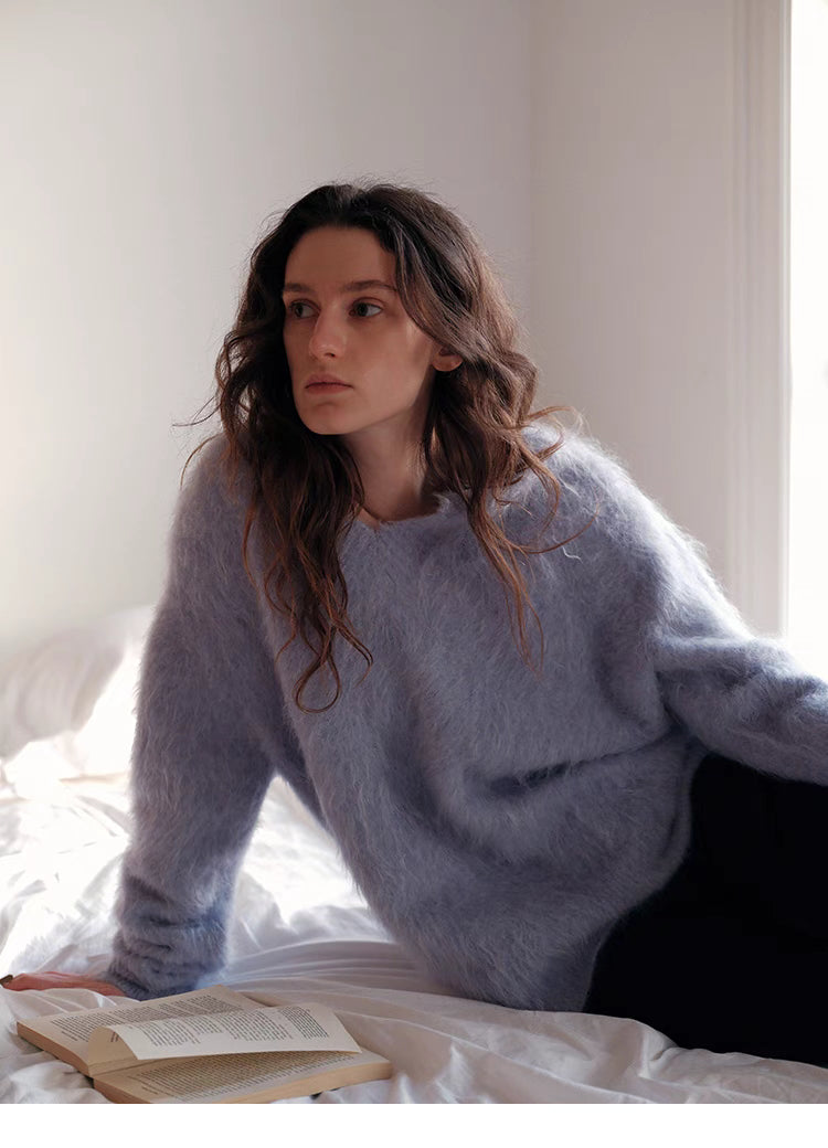 Misty blue wool sweater sitting on bed with relaxed pose highlighting lightweight warmth and soft fabric