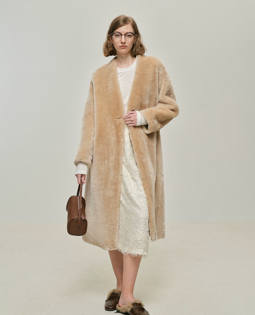 Sand collarless shearling coat worn open, showing straight below-knee length
