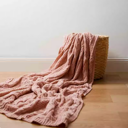 Pink braided knit oversized throw blanket draped from woven basket onto wooden floor showing soft texture and cable knit pattern