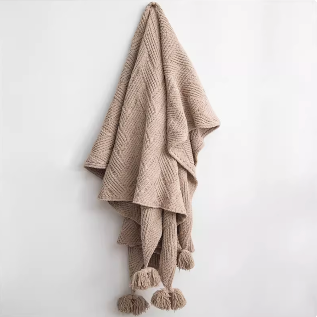 Almond chenille cable knit throw blanket hanging on wall with pom pom edges showing warm neutral tone and soft texture