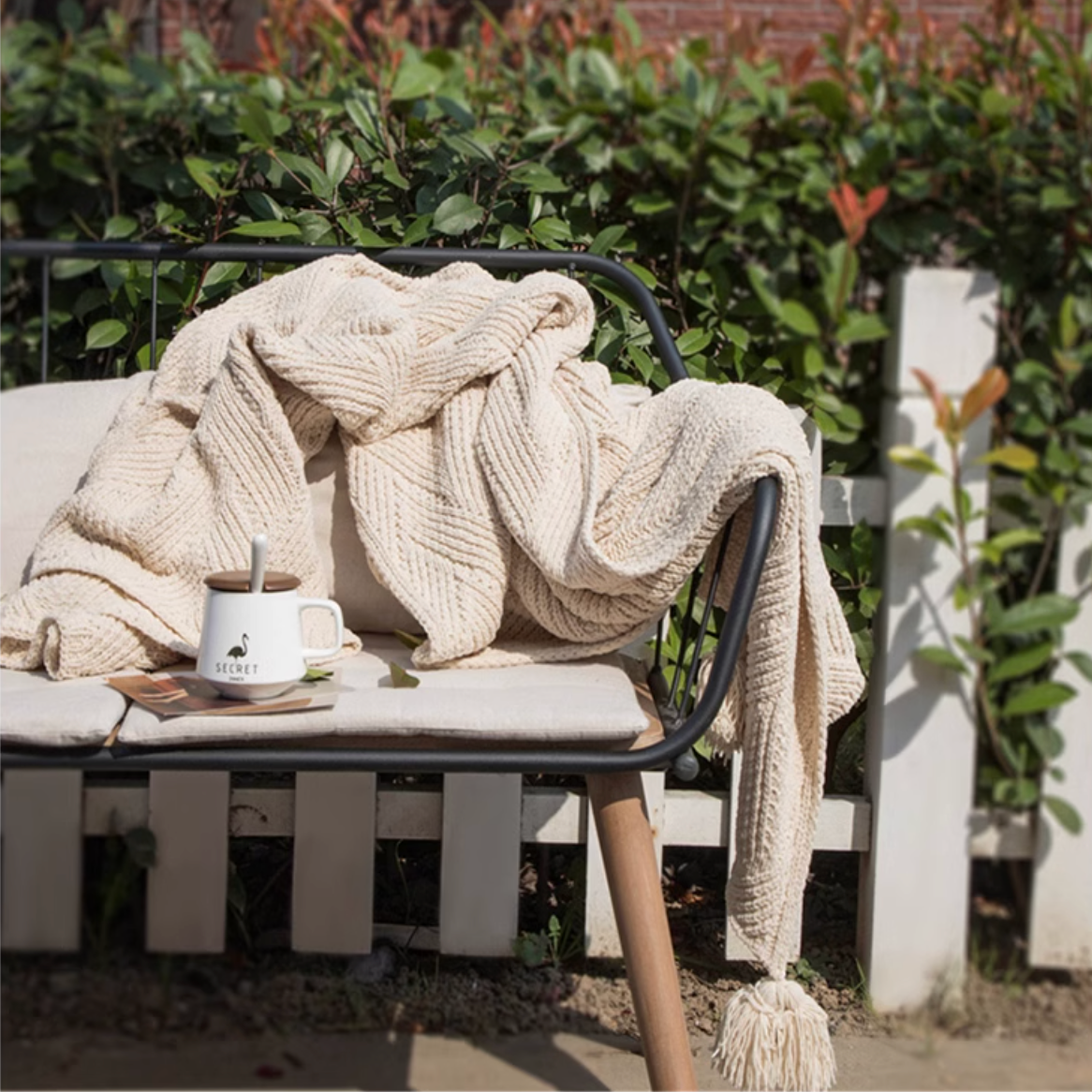 Beige chenille cable knit throw blanket draped over outdoor bench with cup and greenery creating a cozy relaxing setting