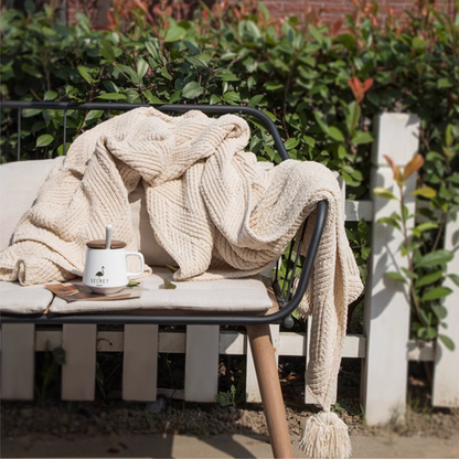 Beige chenille cable knit throw blanket draped over outdoor bench with cup and greenery creating a cozy relaxing setting