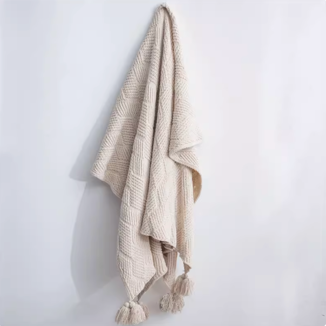 Beige chenille cable knit throw blanket hanging on wall with pom pom edges showing soft textured knit design