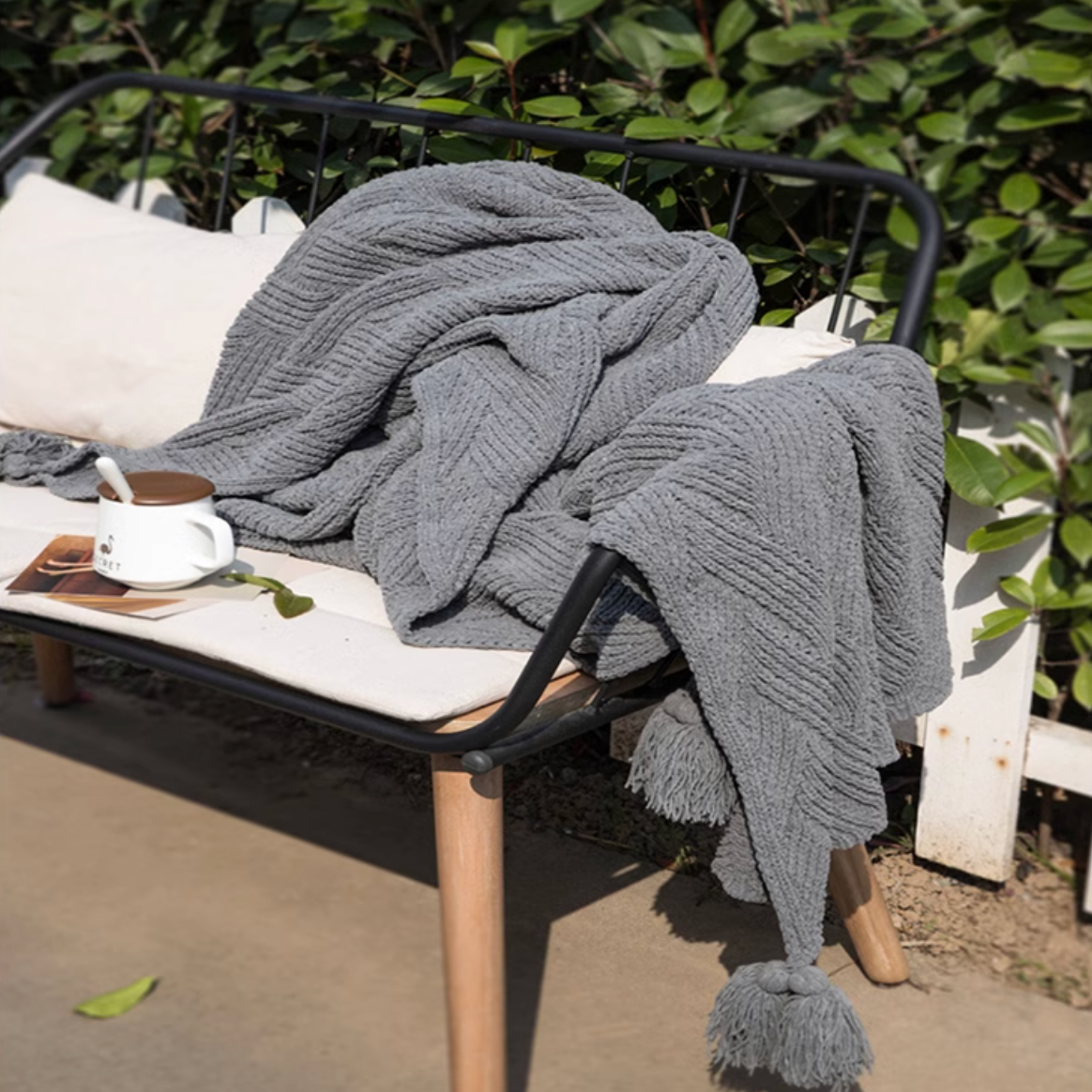 Gray chenille cable knit throw blanket draped over outdoor bench with cup and greenery creating a relaxed cozy environment