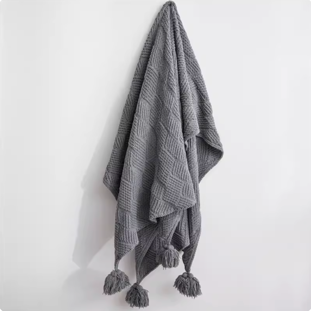 Gray chenille cable knit throw blanket hanging on wall with pom pom edges showing modern neutral tone and soft texture