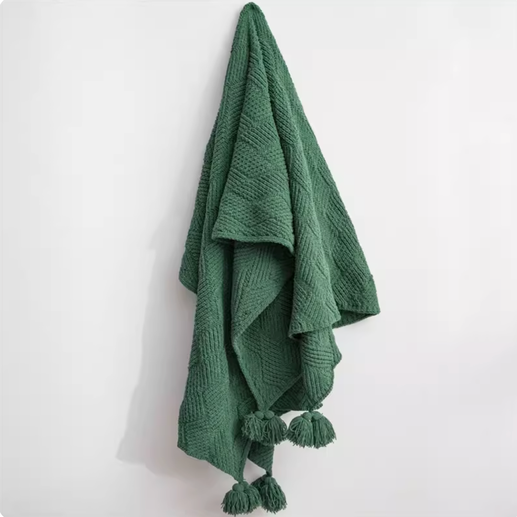 Green chenille cable knit throw blanket hanging on wall with pom pom edges displaying textured knit and vibrant color