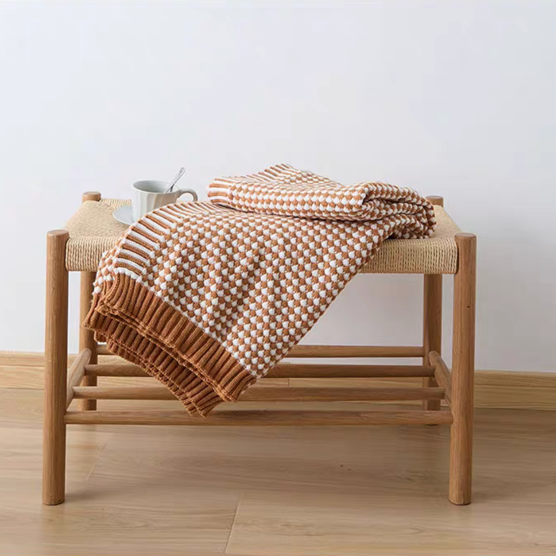 Dark yellow checkered acrylic knit throw blanket folded on a wooden bench with cup, cozy modern home decor styling