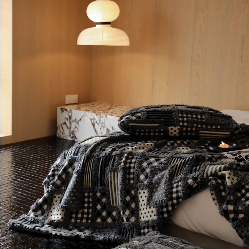 Gray checked patchwork throw blanket draped across bed in a warm bedroom setting with wooden wall and soft bedside lamp lighting