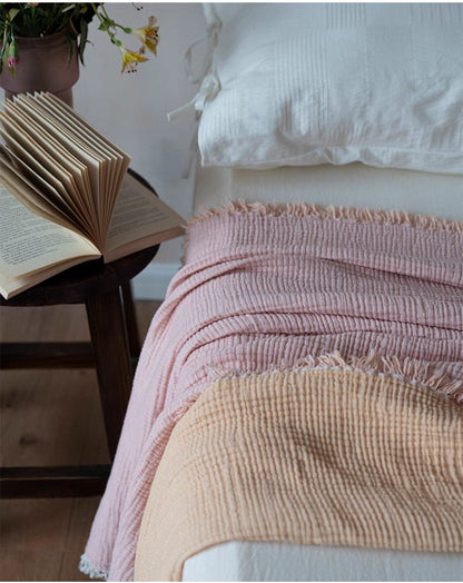 Folded orange pink cotton throw blanket on bed beside nightstand showing breathable gauze weave