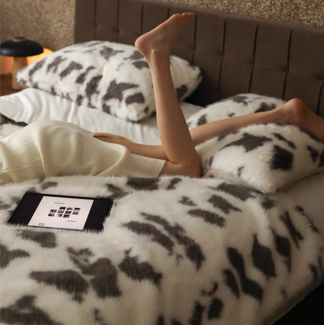 Cow cream faux fur throw blanket on bed with soft plush texture and model relaxing in cozy bedroom setting