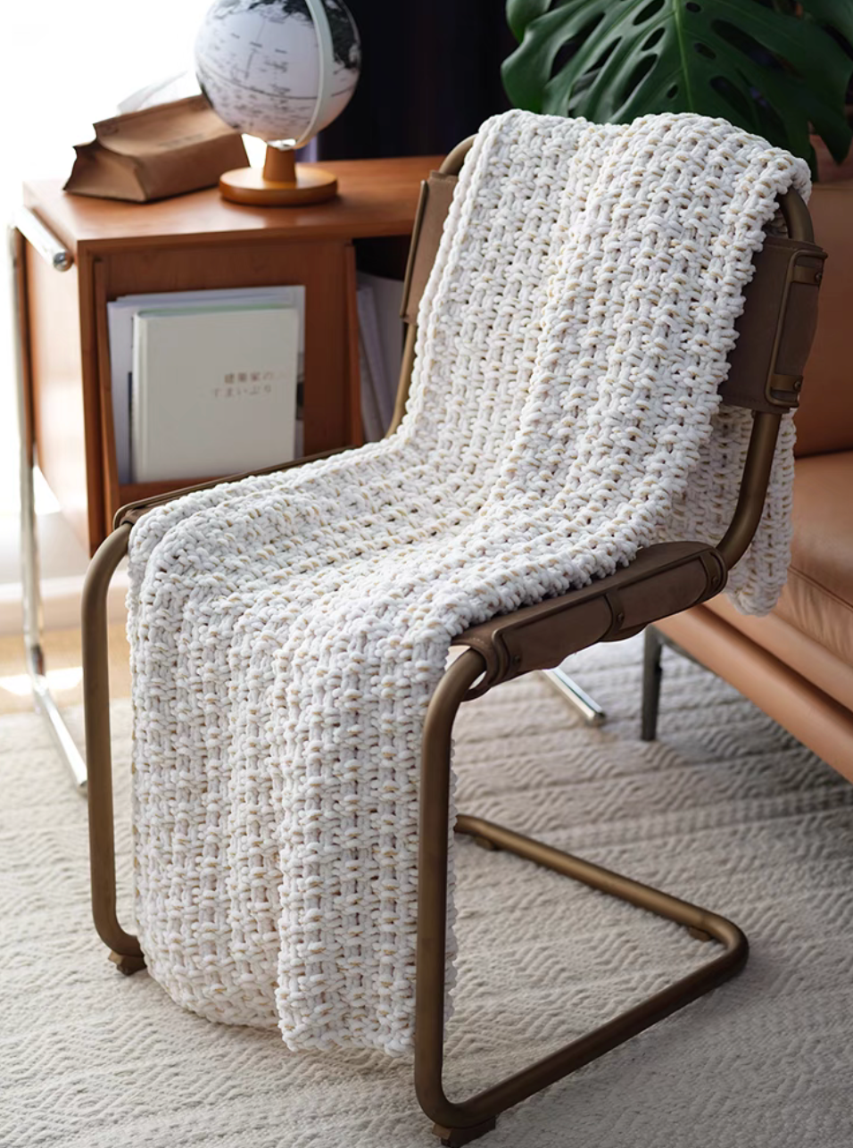 White chunky knit throw blanket draped over a modern chair beside a wooden side table in a cozy room