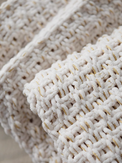 Close-up of folded white chunky knit throw blanket highlighting thick weave, soft texture, and gold thread accents