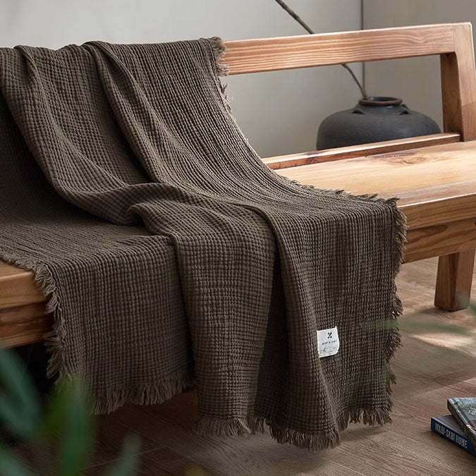 Grey green organic cotton waffle textured fringe throw blanket draped across wooden bench in modern interior.