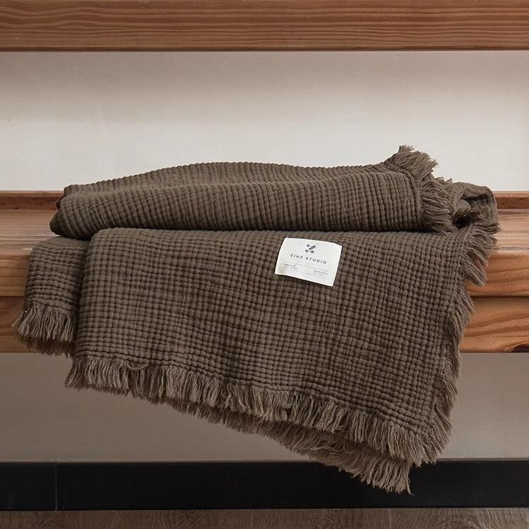 Grey green waffle textured organic cotton throw blanket folded neatly on wooden bench with soft fringe trim.
