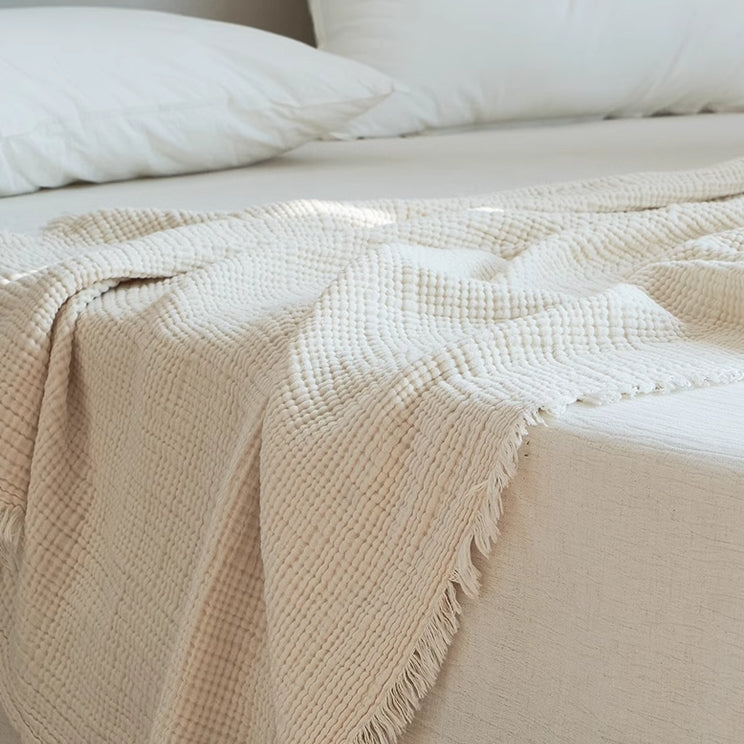 Ivory white organic cotton waffle gauze throw blanket styled across a bed with soft textured weave and raw fringe edges.