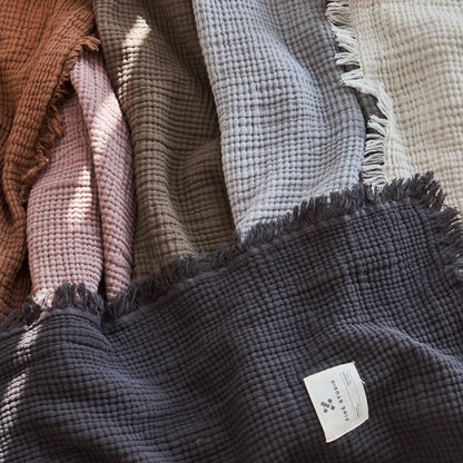 Multiple color organic cotton waffle textured fringe throw blankets displayed together showing caramel, purple, grey and ivory options.