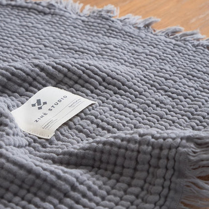 Smoky blue waffle textured organic cotton throw blanket close-up showing breathable gauze weave and fringe trim.